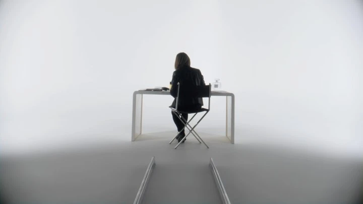 Back view of woman in black jacket sitting at white table with beauty products in bright studio setting with simple metal accents.