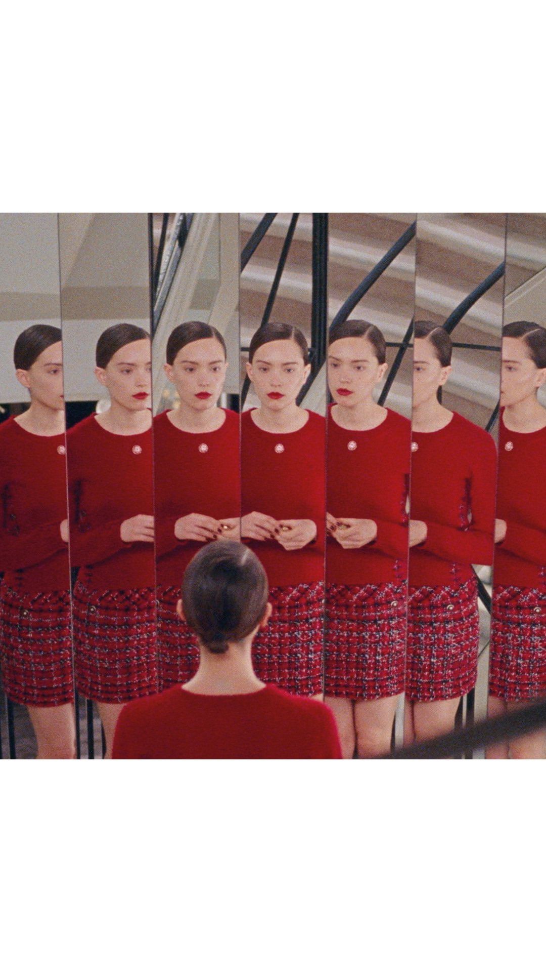 Fashion shot of a woman in a red sweater and tweed skirt facing a mirrored wall, creating multiple reflections of the same person.