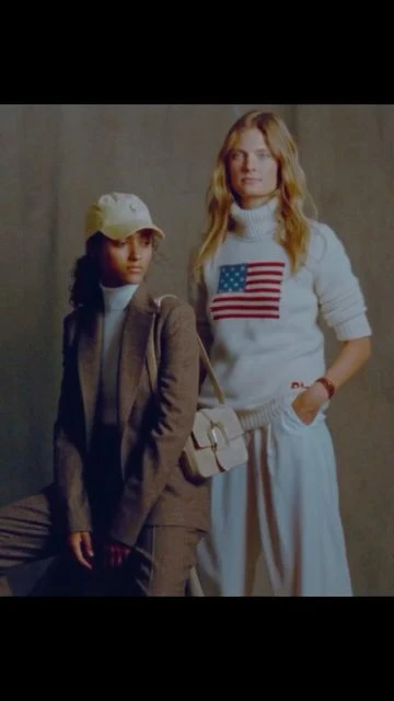 Two fashion models in a studio setting; one wears a tan suit and baseball cap, the other a white sweater with an American flag.