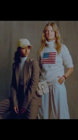 Two fashion models in a studio setting; one wears a tan suit and baseball cap, the other a white sweater with an American flag.
