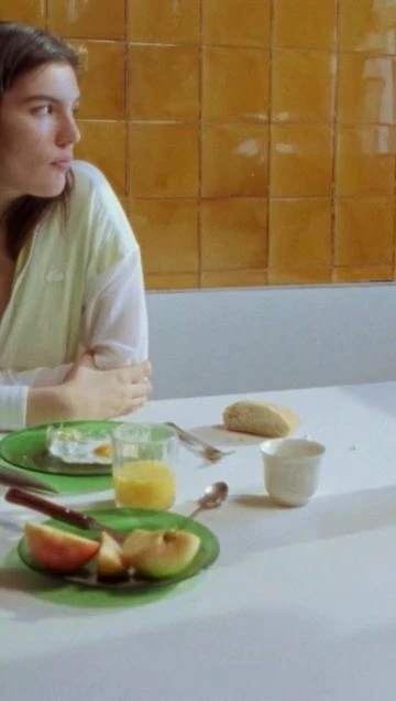 Young woman in a light shirt sits at a table with breakfast foods including sliced apples, juice, bread, and a fried egg on tiles.