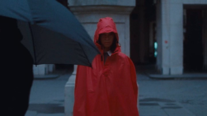 Woman in a hooded red poncho standing outside in an urban setting, partially obscured by a person holding an umbrella.
