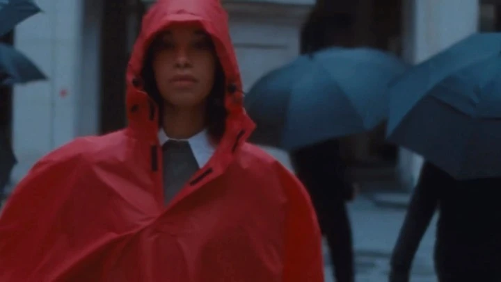 Young woman in a red hooded raincoat walks in an urban environment with people holding umbrellas in the background.