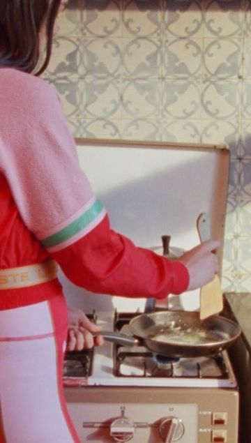 Woman in a pink and red sweatsuit cooks food in a silver frying pan on a white and brown oven in an indoor setting.