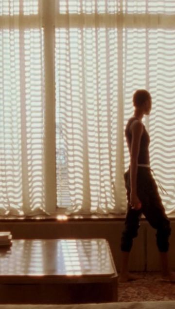 Woman in a tank top and sweatpants stands by a window with wavy patterned curtains in a dimly lit room with a wooden table.