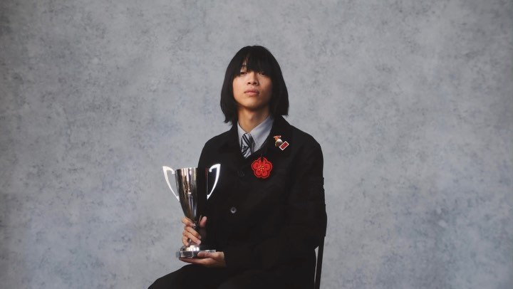 Portrait of an Asian male seated holding a trophy. He wears a black jacket with a flower pin, with a grey textured background.