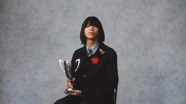 Portrait of an Asian male seated holding a trophy. He wears a black jacket with a flower pin, with a grey textured background.