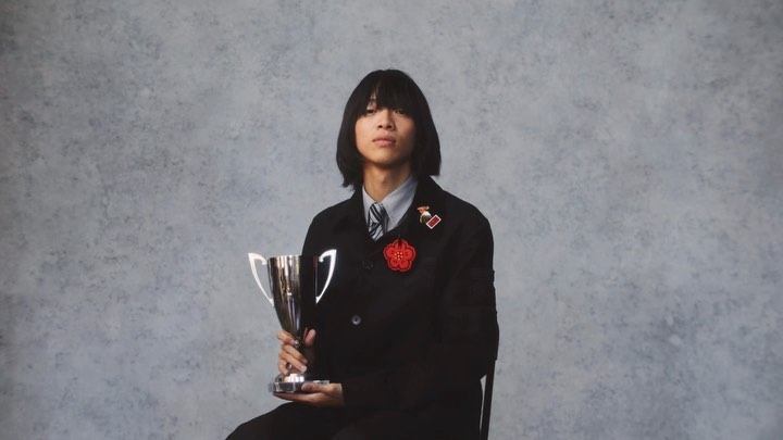 Young adult poses indoors holding a silver trophy wearing dark blazer with red floral pin against a textured mottled grey backdrop.