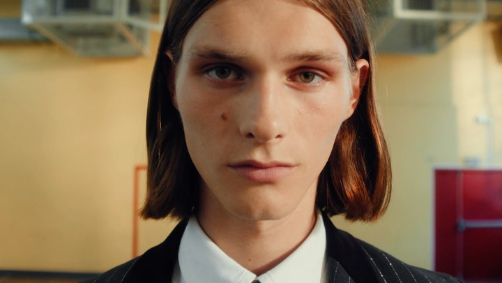 Close-up portrait of a young man with long brown hair wearing a white collared shirt and dark suit jacket.