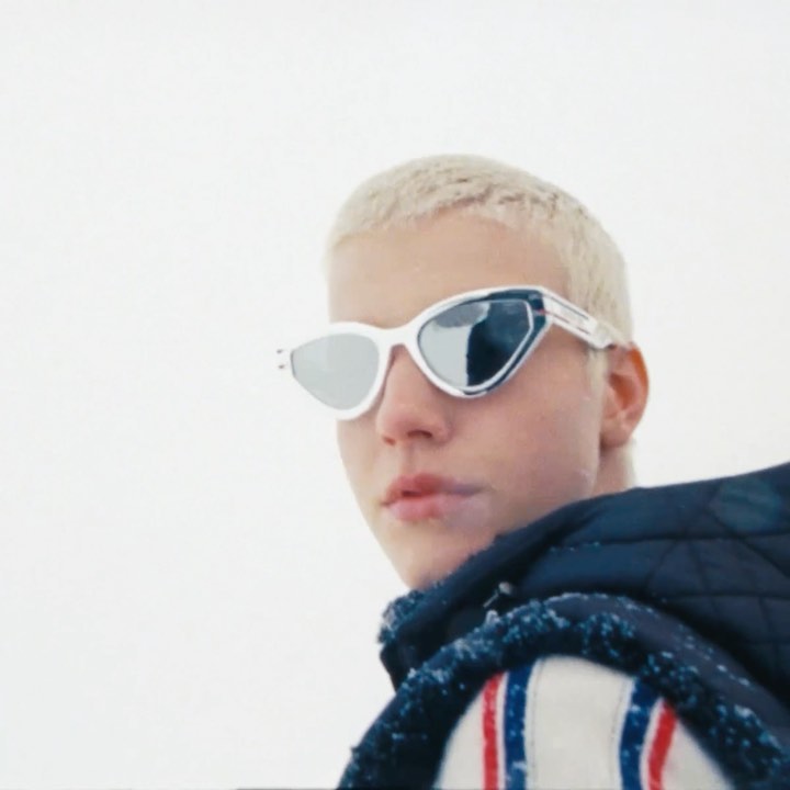 Close-up of a young adult with platinum blonde hair wearing futuristic white sunglasses with silver mirrored lenses, neutral expression.