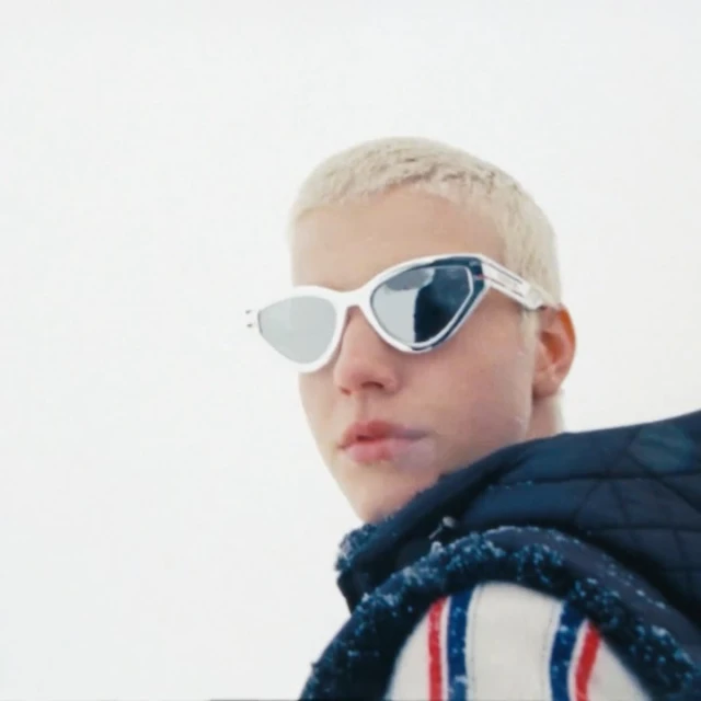 Close-up of a young adult with platinum blonde hair wearing futuristic white sunglasses with silver mirrored lenses, neutral expression.