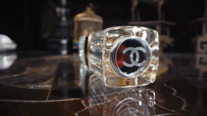 Close-up of a crystal perfume bottle with a logo on a shiny surface in a dimly lit setting, with a birdcage in the background.