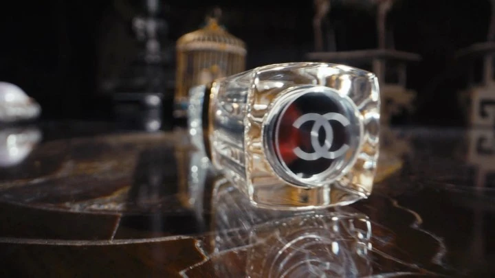 Close-up of a crystal perfume bottle with a logo on a shiny surface in a dimly lit setting, with a birdcage in the background.
