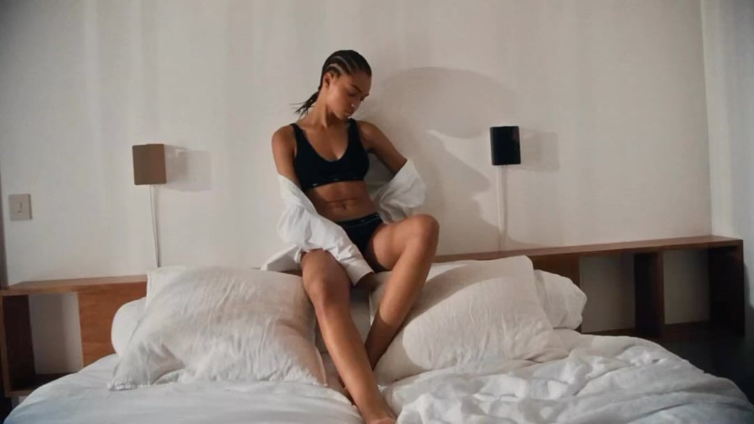 An adult woman with cornrow braids sitting on an unmade bed in a black lingerie set and a white button-down shirt in a bedroom.