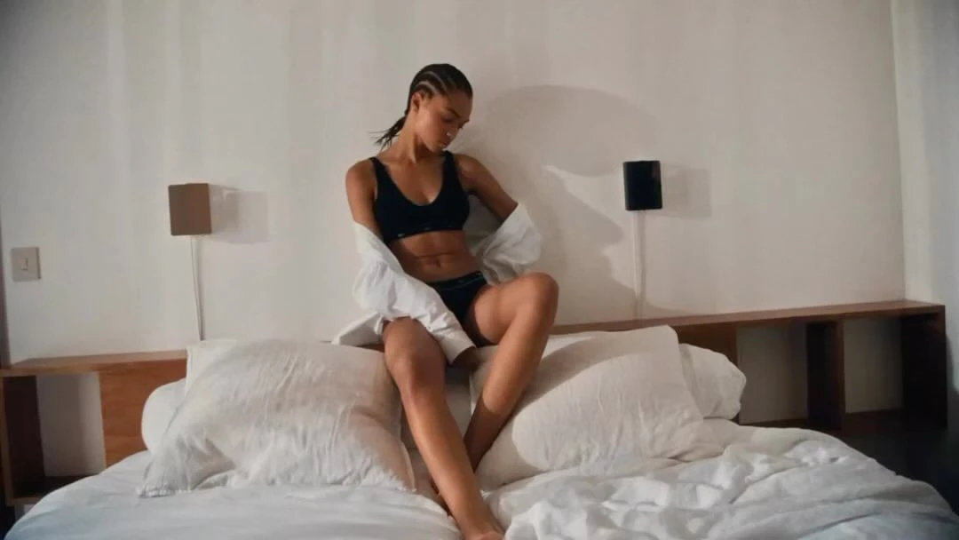 An adult woman with cornrow braids sitting on an unmade bed in a black lingerie set and a white button-down shirt in a bedroom.