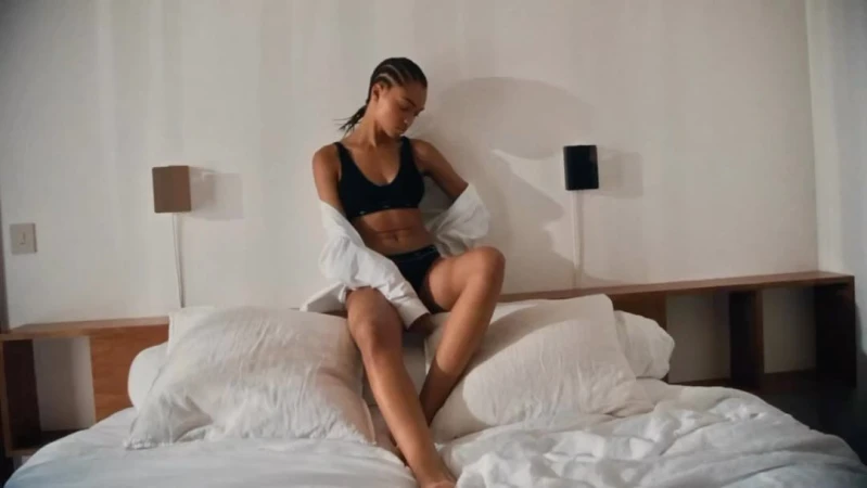 An adult woman with cornrow braids sitting on an unmade bed in a black lingerie set and a white button-down shirt in a bedroom.