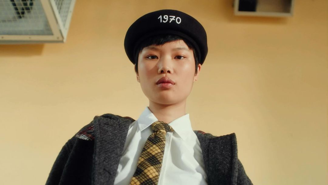 Portrait of an Asian woman wearing a beret and tie with a neutral expression in front of a muted yellow background in a studio.