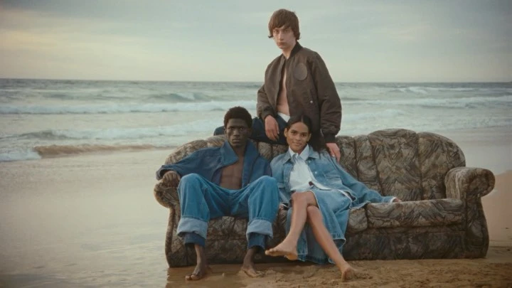Three models sit on a patterned sofa on a sandy beach with ocean waves in the background, wearing denim and jacket in an outdoor fashion shoot.