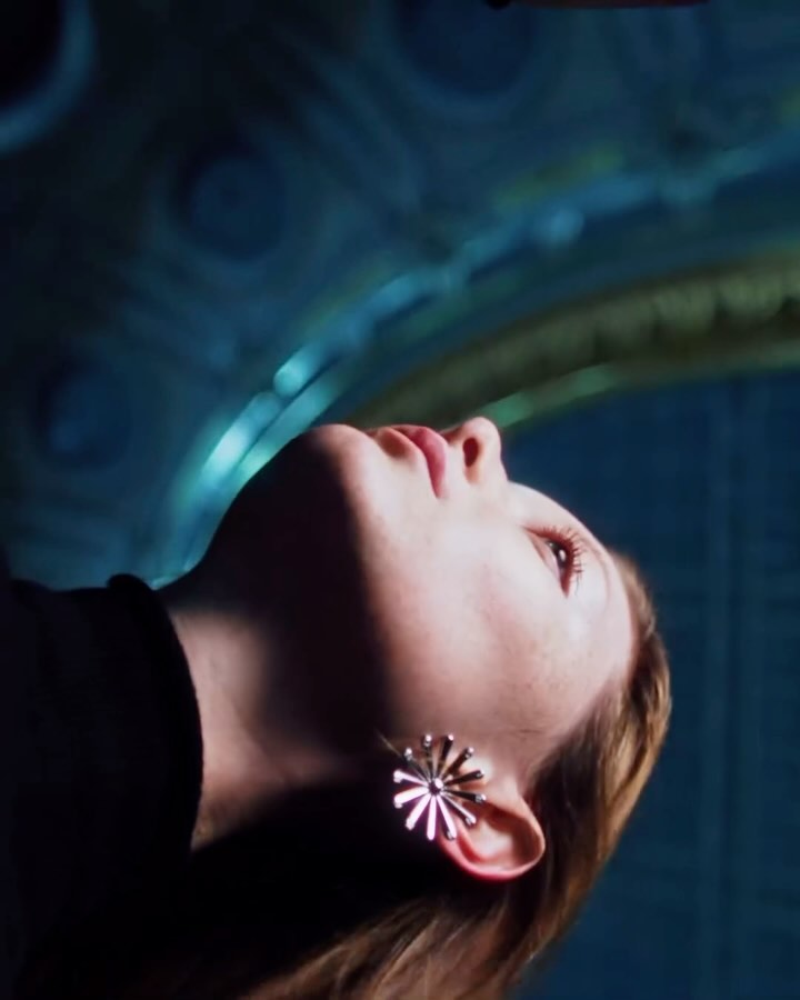 Low-angle shot of a young woman with blonde hair and flower earrings looking up, illuminated by blue light against an ornate patterned backdrop.
