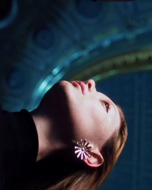 Low-angle shot of a young woman with blonde hair and flower earrings looking up, illuminated by blue light against an ornate patterned backdrop.