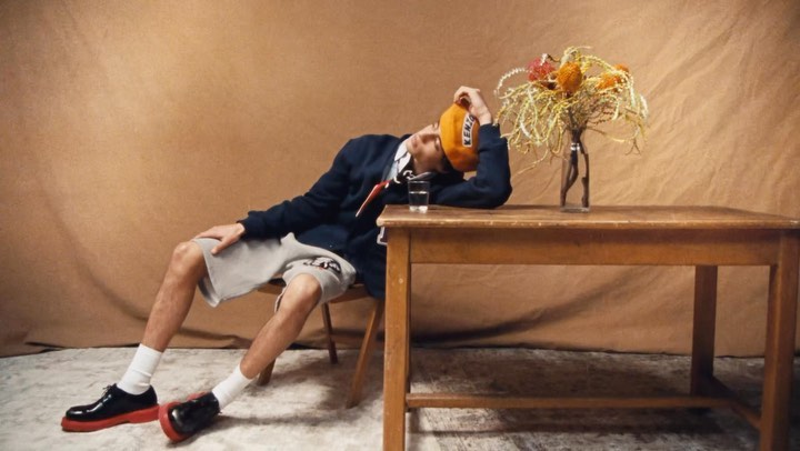 Adult male in casual collegiate style rests head on wooden table next to glass of water and flowers in front of tan backdrop.