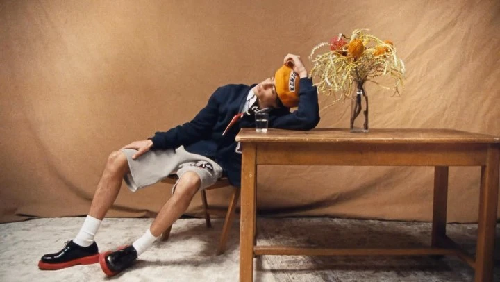 Adult male in casual collegiate style rests head on wooden table next to glass of water and flowers in front of tan backdrop.