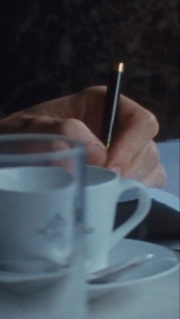 Close-up of a hand writing with a black pen on paper, with a white teacup and saucer set in the foreground, and a dark background.