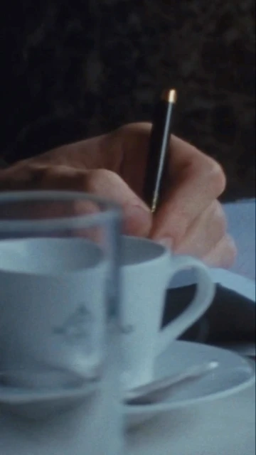 Close-up of a hand writing with a black pen on paper, with a white teacup and saucer set in the foreground, and a dark background.