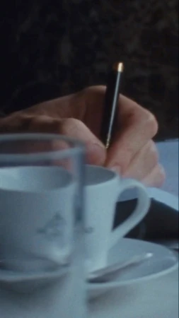 Close-up of a hand writing with a black pen on paper, with a white teacup and saucer set in the foreground, and a dark background.