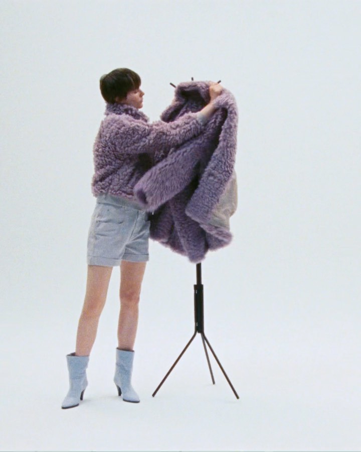 Woman in a purple, textured jacket and jean shorts posing with a purple jacket on a clothing rack in a bright, neutral studio.