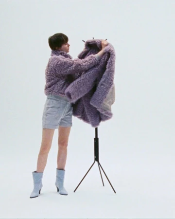 Woman in a purple, textured jacket and jean shorts posing with a purple jacket on a clothing rack in a bright, neutral studio.