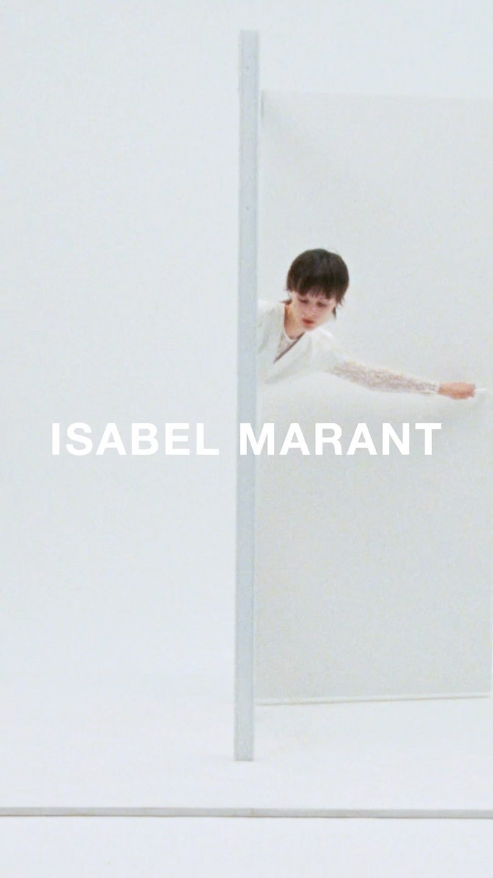 A fair-skinned young adult poses in front of an off-white studio backdrop, wearing a white lace-sleeved top, peering through a partition.