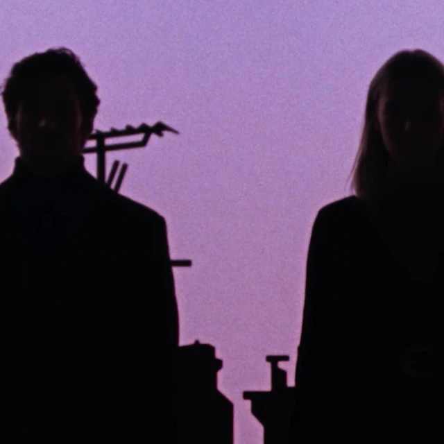 Silhouetted couple against a purple sky, architectural details visible on the left side of the frame, creating a stark contrast.
