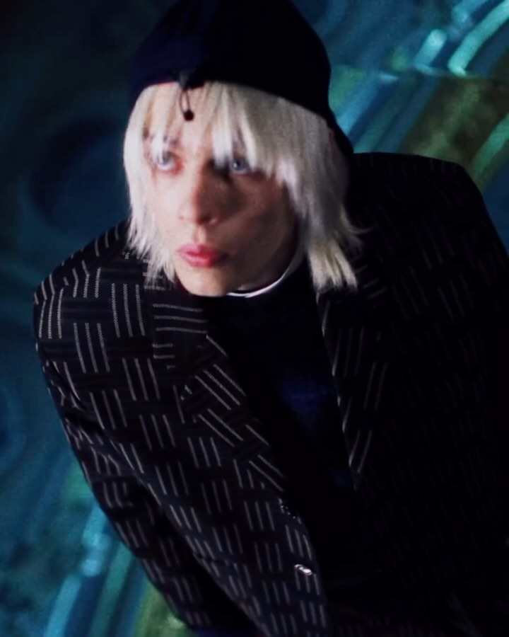 Close-up of a person with platinum blonde hair wearing a blue cap and patterned blazer, looking intently to the side, against a blue background.