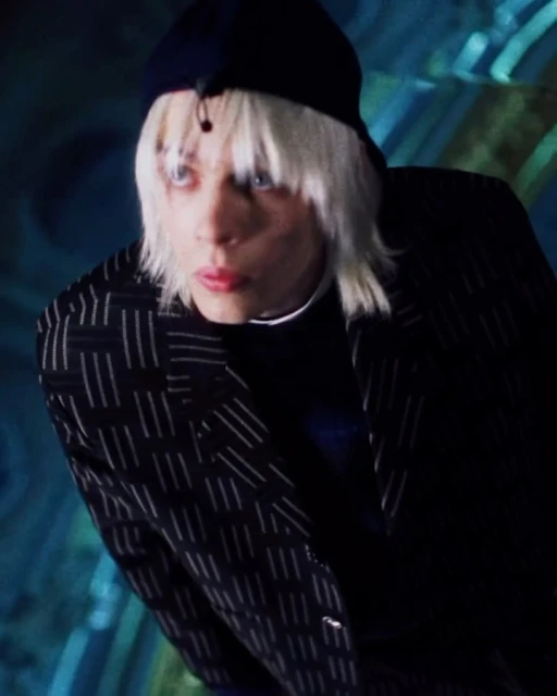 Close-up of a person with platinum blonde hair wearing a blue cap and patterned blazer, looking intently to the side, against a blue background.