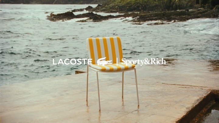 Coastal scene featuring a Lacoste branded white chair with yellow and white stripes, set against a rocky shore and ocean backdrop.