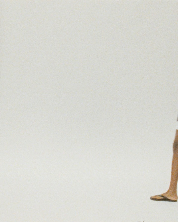 Partial view of an adult with dark hair, wearing shorts and flip-flops, stands against a plain white background, creating a minimalistic fashion shot.