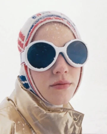 Close-up portrait of a woman wearing large round white sunglasses, a patterned winter cap, and a shiny metallic gold jacket in the snow.