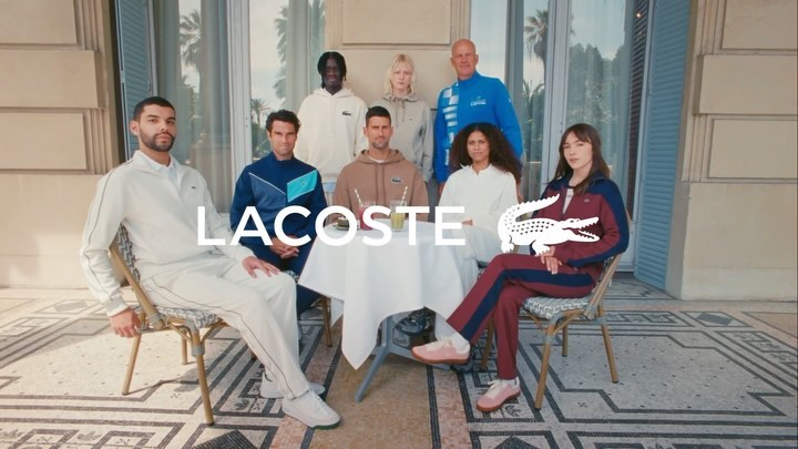 Group of fashion models wearing Lacoste apparel, sitting at a table outdoors on a sunny day with the brand logo in the foreground.