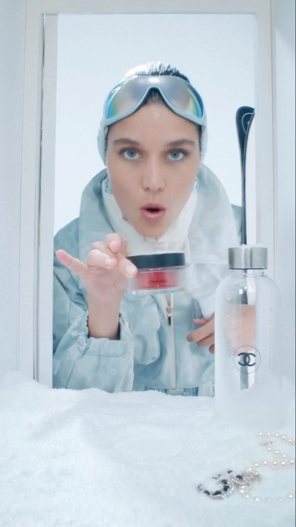 A woman in ski goggles and winter clothing presents a jar of red cream in a bright, snowy setting, with a bottle of Chanel fragrance.