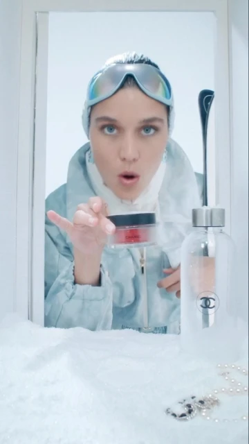 A woman in ski goggles and winter clothing presents a jar of red cream in a bright, snowy setting, with a bottle of Chanel fragrance.