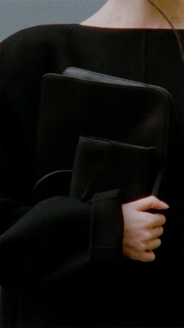 Mid-shot of an adult female holding a black portfolio, wearing a dark top with sleeves, showing hands and minimalist fashion.