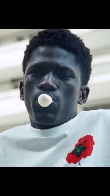 Close-up of a young Black man blowing a bubble with gum; he wears a white t-shirt with a red poppy design and stares straight ahead.