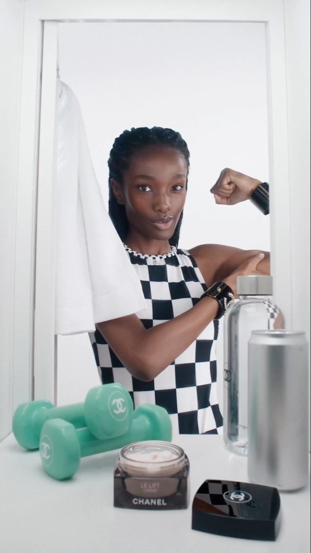 Fit Black woman in sportswear flexing muscle next to dumbbells and beauty products inside of doorway against white backdrop.