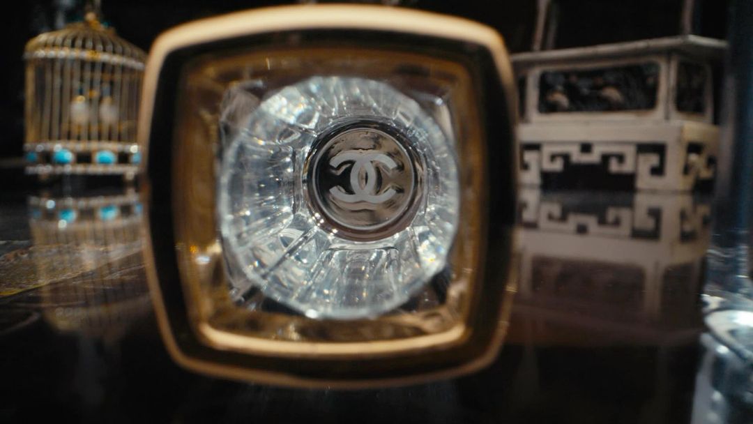 Close-up of a clear glass bottle with a gold rim and the Chanel logo visible, a decorative birdcage and table in the background.