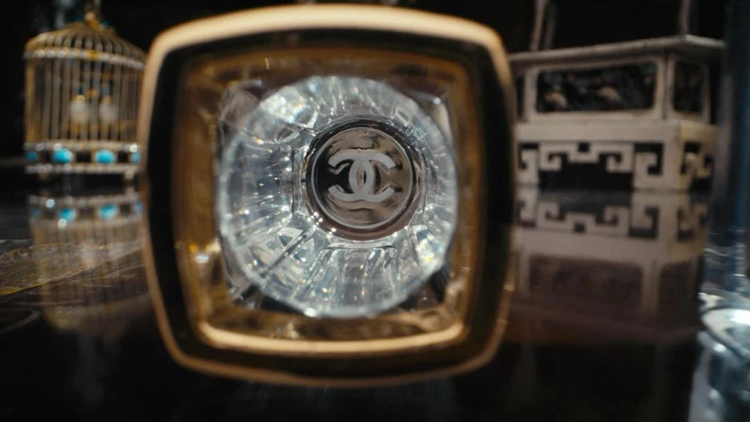 Close-up of a clear glass bottle with a gold rim and the Chanel logo visible, a decorative birdcage and table in the background.