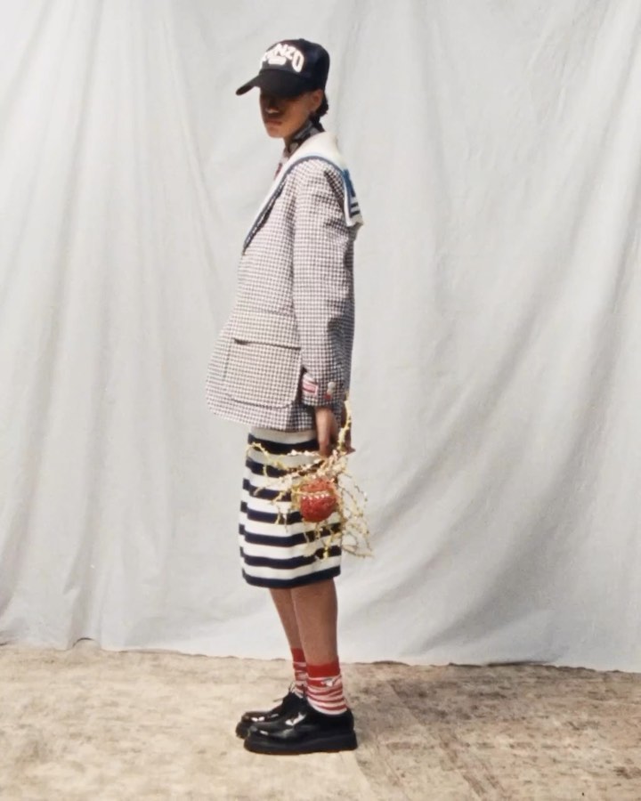 Young adult poses in studio, wearing a blazer, striped skirt, hat, socks and patent leather shoes against a white backdrop.