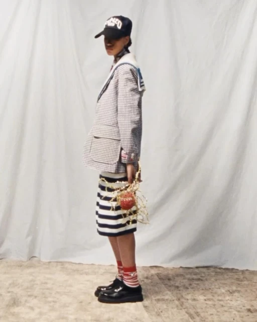 Young adult poses in studio, wearing a blazer, striped skirt, hat, socks and patent leather shoes against a white backdrop.