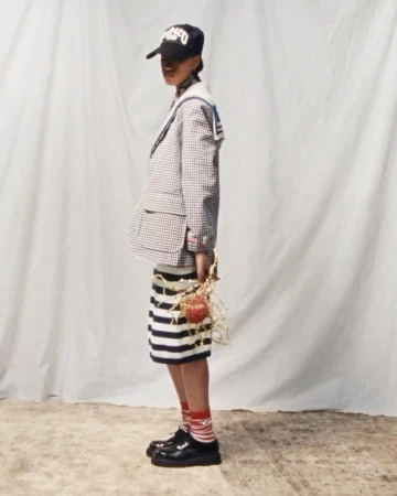 Young adult poses in studio, wearing a blazer, striped skirt, hat, socks and patent leather shoes against a white backdrop.