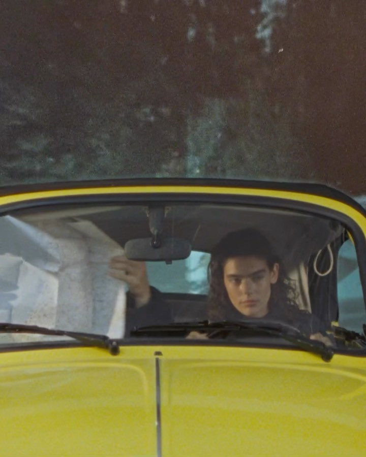 A young adult with long curly hair is seen in a yellow vintage car, looking at a map. The background shows a blurred landscape.